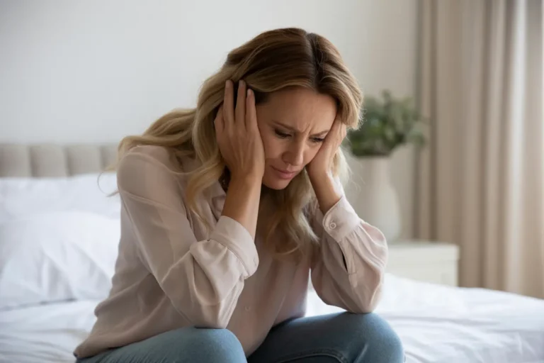 A woman on a bed holds her head in her hands, appearing deep in negative thought