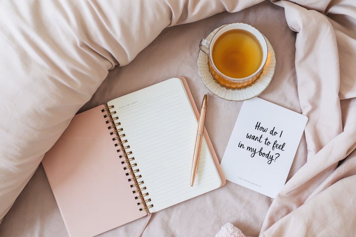 A blush journal, gold pen, herbal tea, and a note that asks “How Do I Want to Feel in My Body?” rest on soft pastel bedding, creating a peaceful scene for clarity.